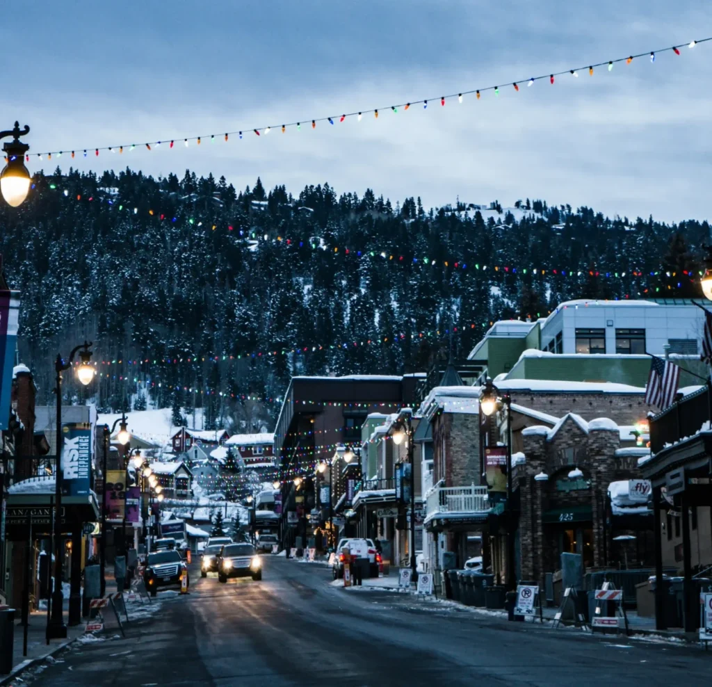 Snowy street with decorated buildings, cars, and colorful lights at dusk.