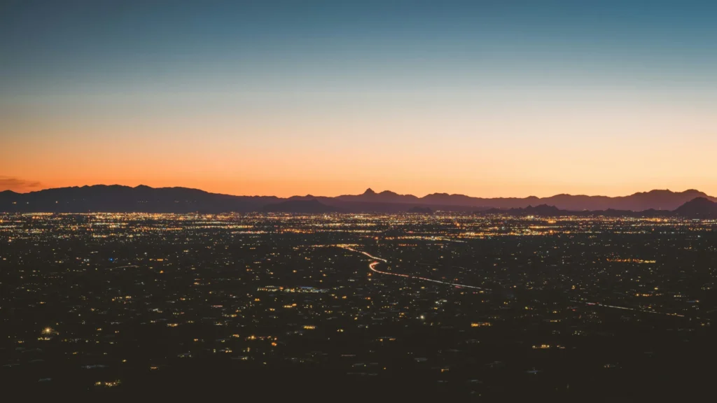 City lights glow at dusk beneath distant mountains and an orange sunset sky.